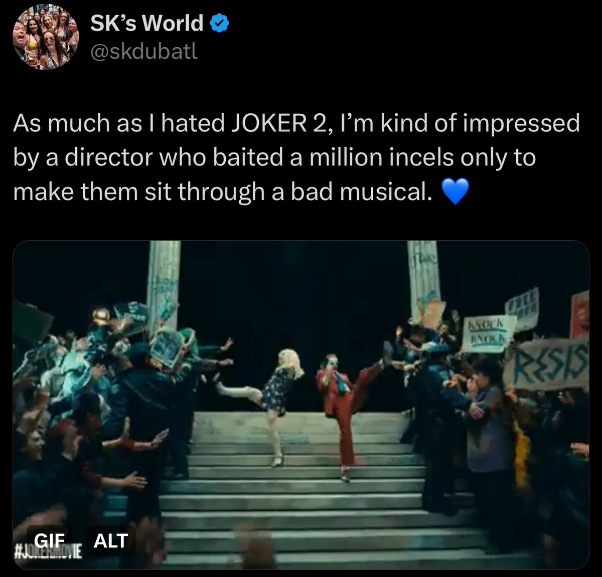 A screenshot from the movie 'Joker 2' shows two characters dancing on steps while a crowd of people with signs surrounds…