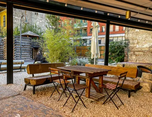 An outdoor patio with wooden tables and benches surrounded by gravel, greenery, and buildings. A person is seated in the…