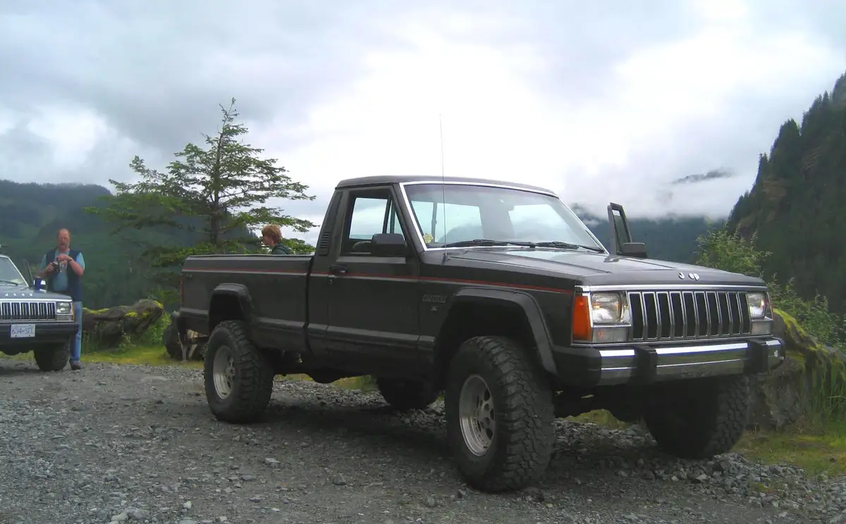 A black, lifted Jeep Comanche pickup truck is parked on a gravel road in a mountainous, forested area under an overcast sky…