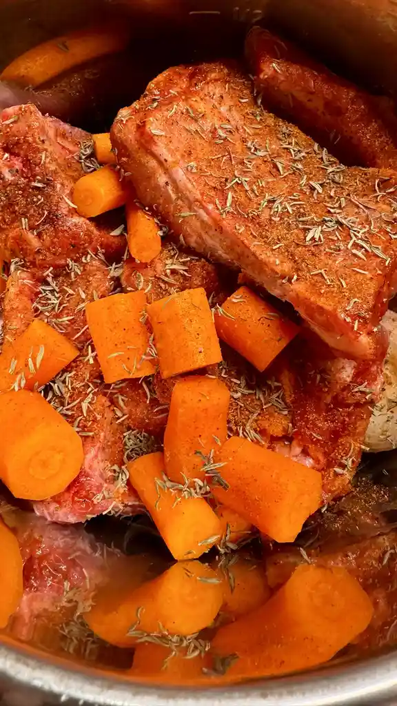 A close-up photo shows raw meat, likely ribs, seasoned with a reddish spice blend and dried herbs, nestled in a slow cooker…