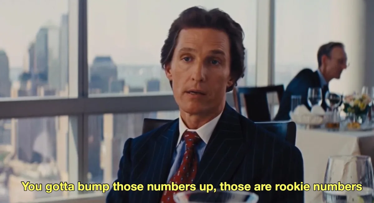 A man in a pinstripe suit and red tie sits at a restaurant table with a city skyline visible through large windows behind…
