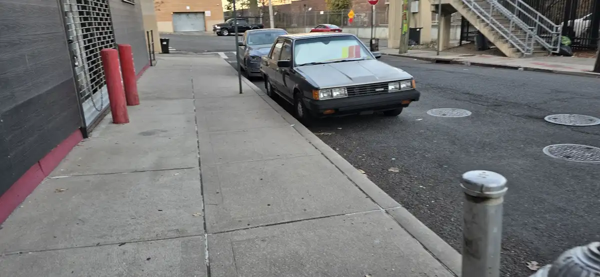 A gray sedan is parked crookedly on a city street, angled sharply into the sidewalk, with a red and yellow 'for sale' sign…