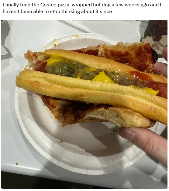 A hand holds a hot dog in a bun that is wrapped in a slice of pizza, topped with yellow mustard, ketchup, and relish. The…