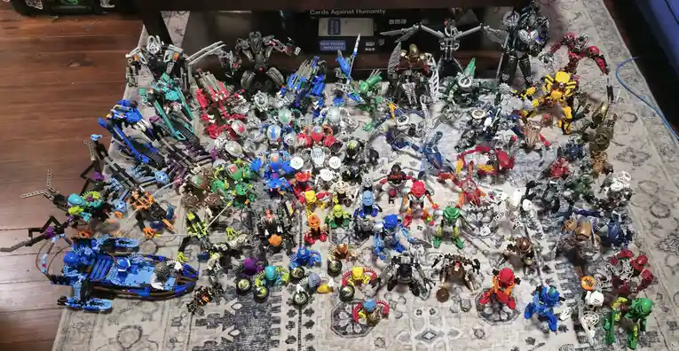 An overhead shot displays a massive collection of Bionicle action figures and builds spread across a patterned rug on a…