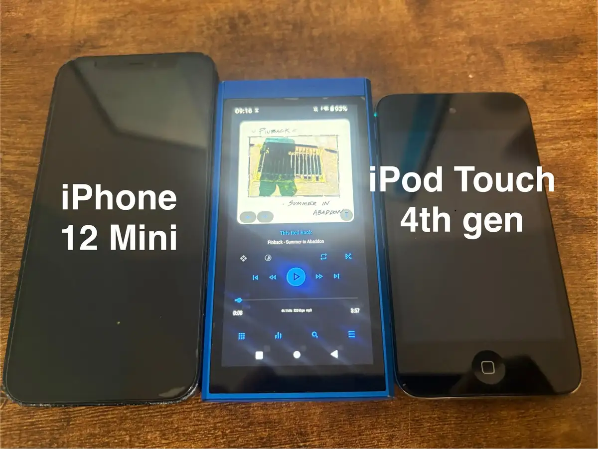 Three devices sit side-by-side on a wooden surface: an iPhone 12 Mini on the left, a blue iPod Touch 4th gen on the right…