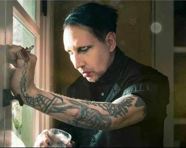 A man with dark hair, heavy makeup, and tattoos on his arm is holding a glass and a key while looking intently at the…