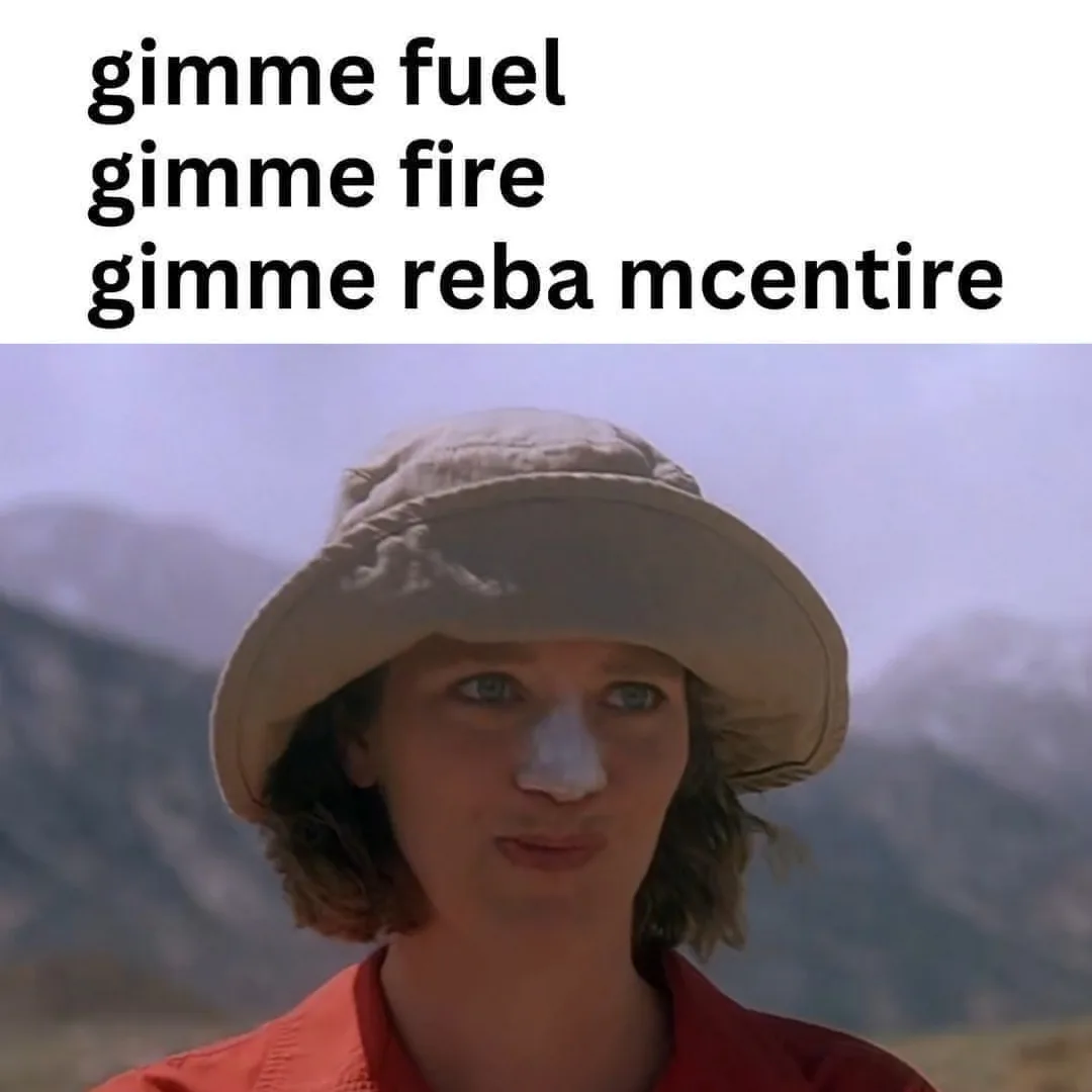 A person wearing a beige bucket hat and red shirt stands outdoors with mountains in the background, looking directly at the…