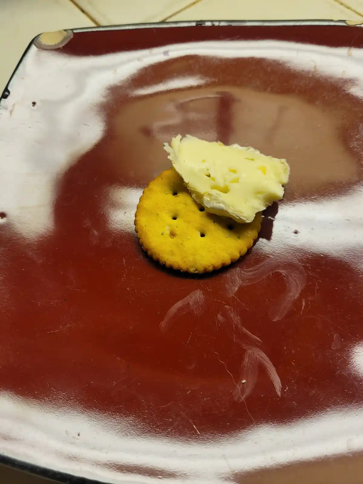 A single yellow saltine cracker with a small, irregularly shaped pat of butter sits centered on a worn, red and white enamel…