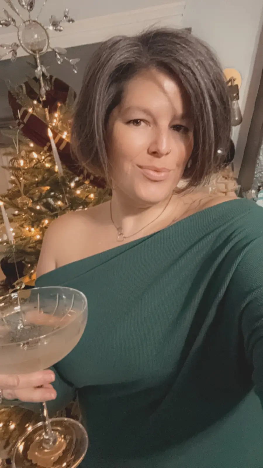A woman with short dark hair wearing a green off-the-shoulder top holds a champagne coupe glass in front of a lit christmas…
