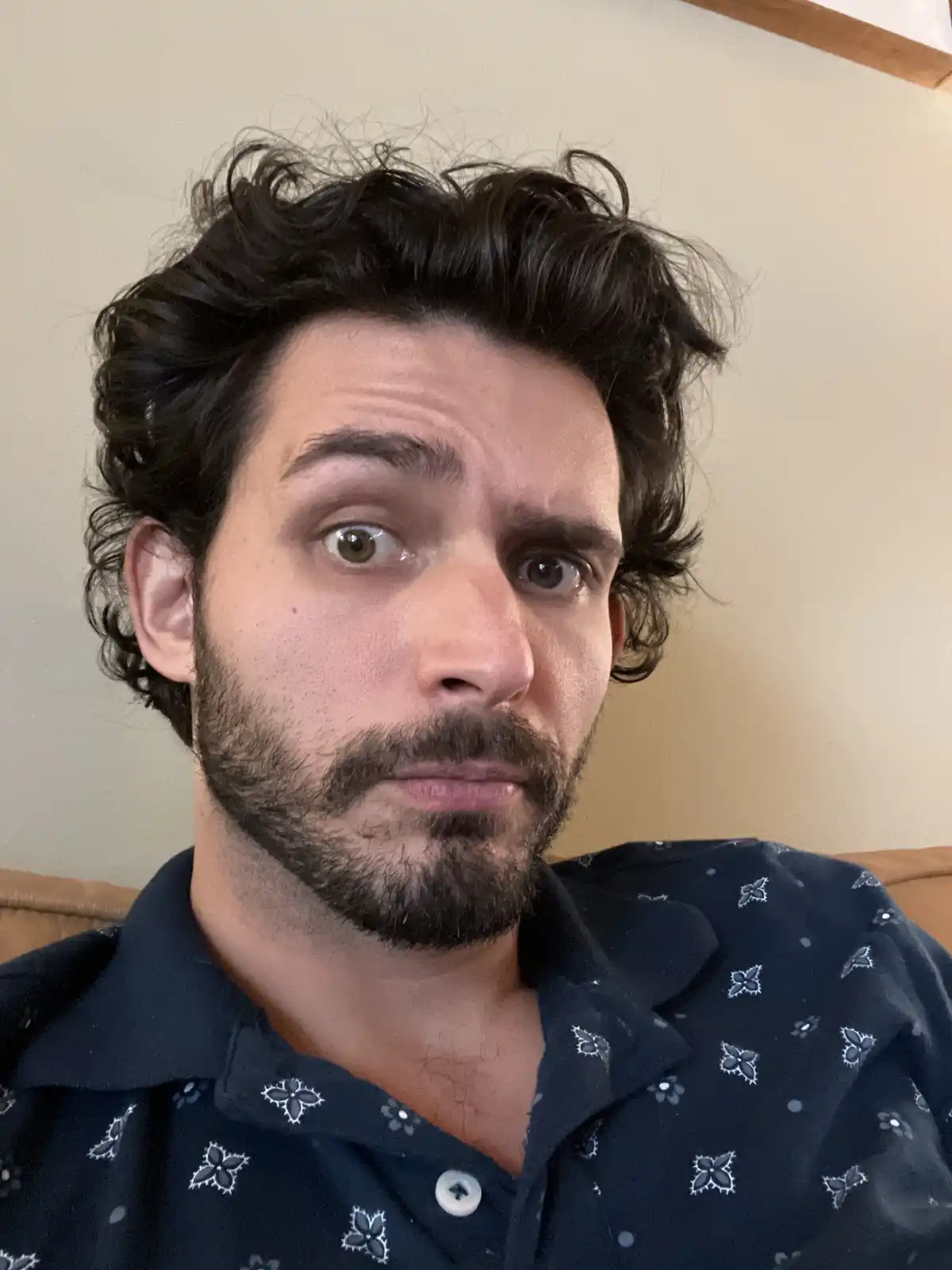 A man with dark curly hair and a beard stares directly at the camera with wide, startled eyes and a slightly downturned…