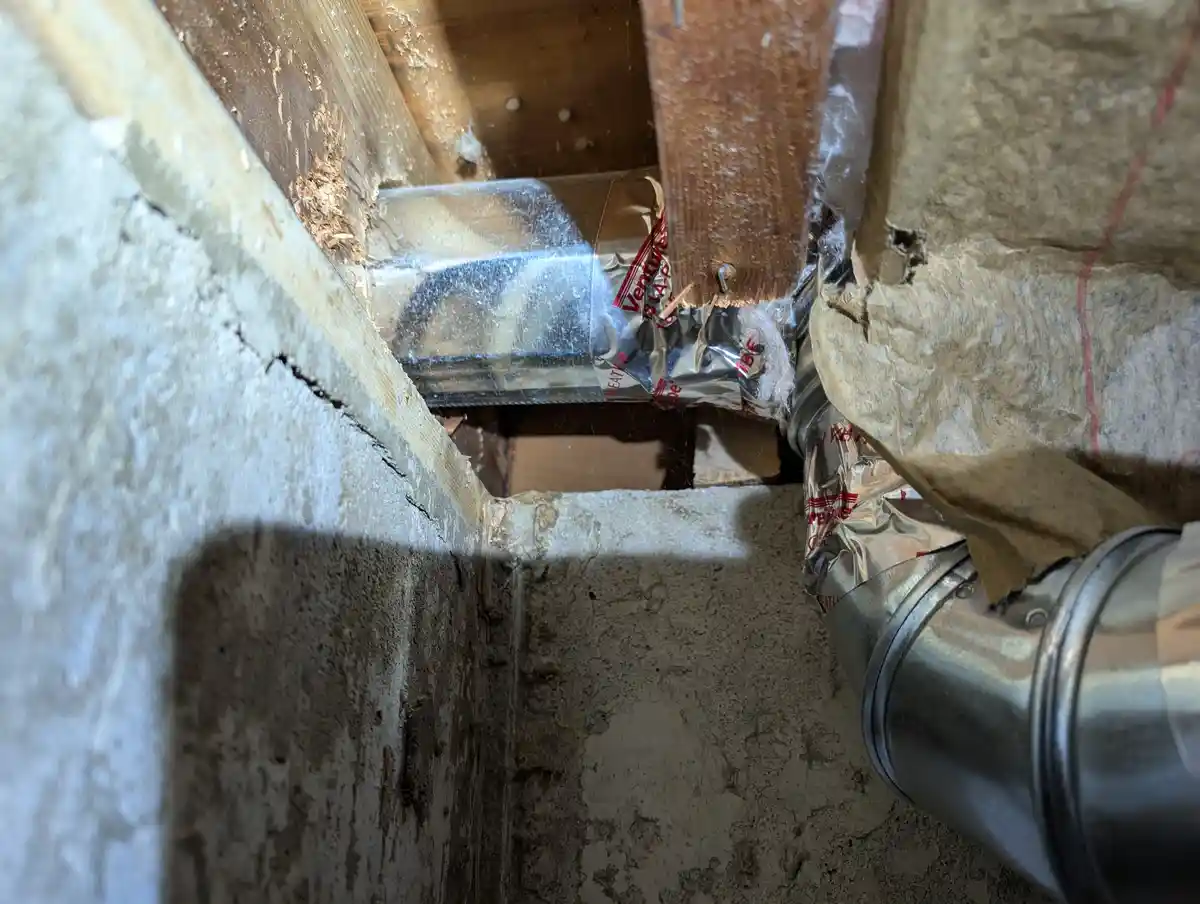 A close-up photo shows a metal ventilation duct running through an attic space, surrounded by wooden beams, insulation, and…