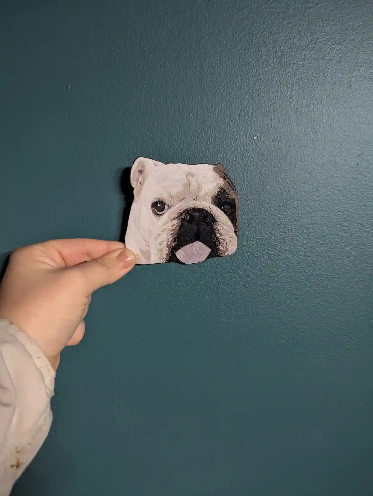 A hand holds up a roughly shaped rock painted to resemble a bulldog's face, featuring white, black, and pinkish tones…