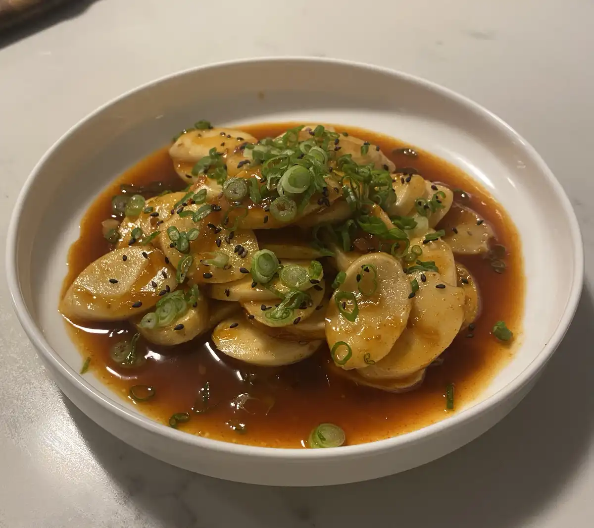 A white bowl contains sliced, saucy food resembling dumplings or potatoes in a reddish-brown sauce, topped with chopped…