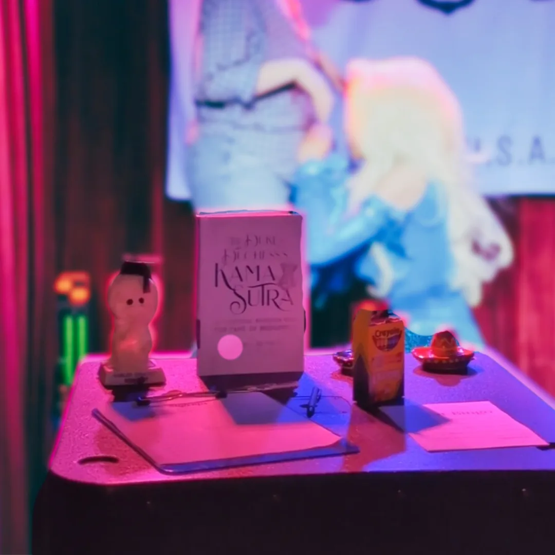 A table under pink and blue neon light holds a book titled 'The Dark Duchess KAMA SUTRA', a small figurine, a box of Crayola…
