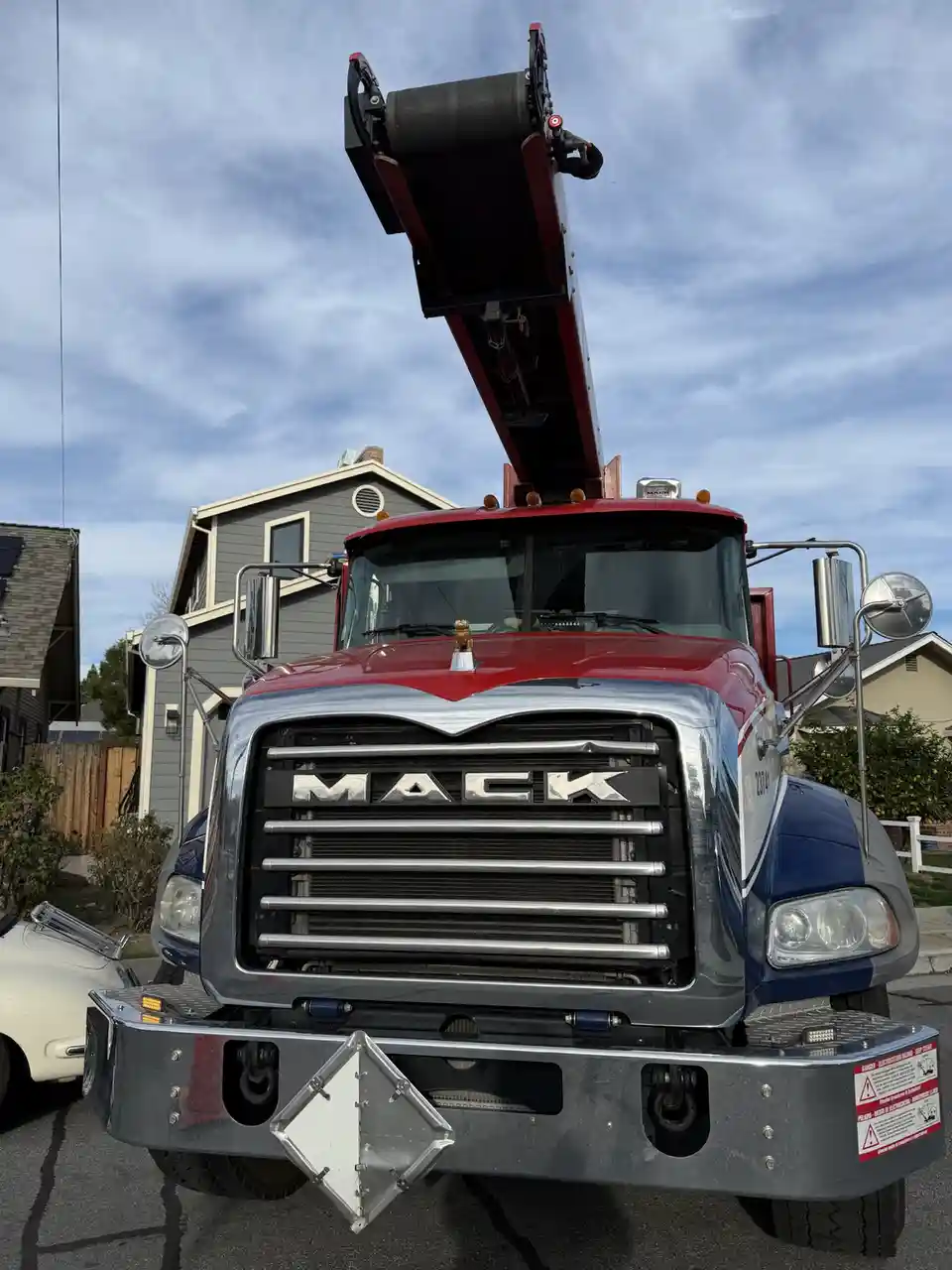 A red and blue Mack truck with a large crane arm extended upwards is parked on a residential street. The truck's chrome…