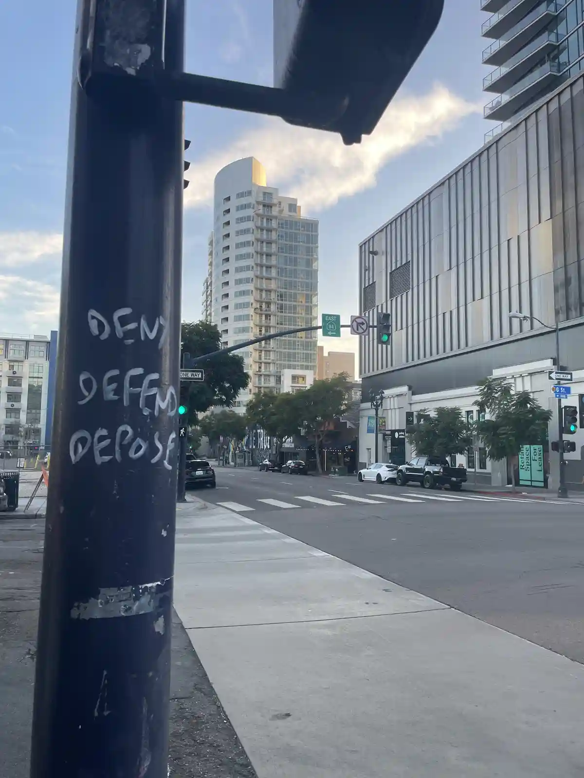 A street view shows a black pole with the words 'DENY', 'DEFEND', and 'DEPOSE' written in white graffiti. The background…