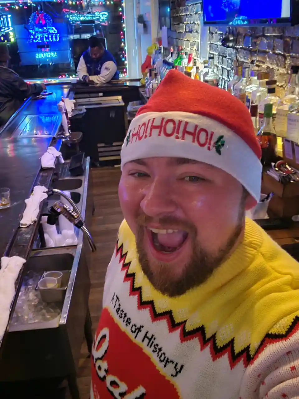 A man in a bar wearing a red and white Santa hat with 'HO! HO! HO!' printed on it and a bright yellow sweater with 'A Taste…