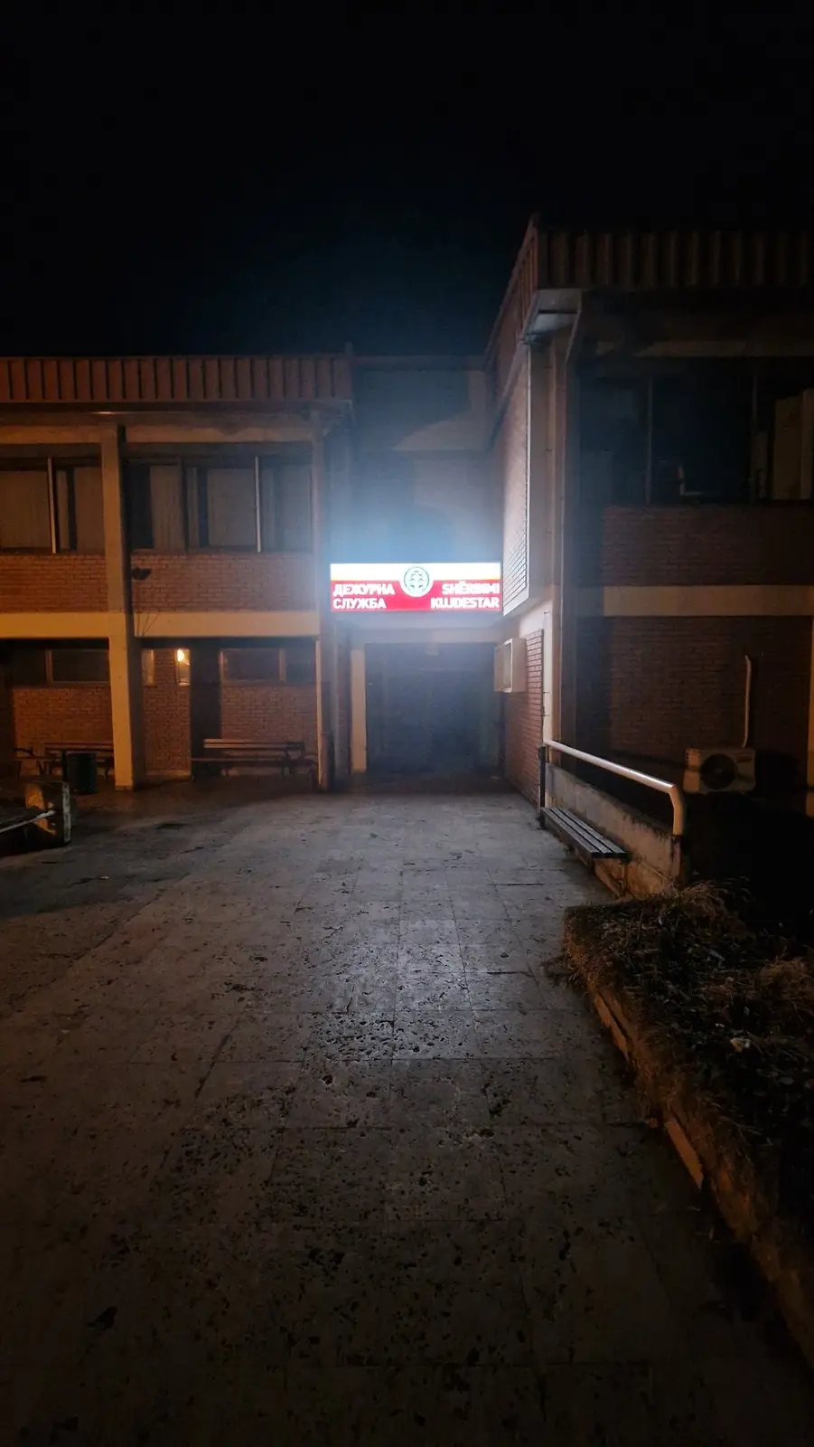 A nighttime photo of a dimly lit, somewhat rundown brick building with a brightly illuminated red sign above an entrance…