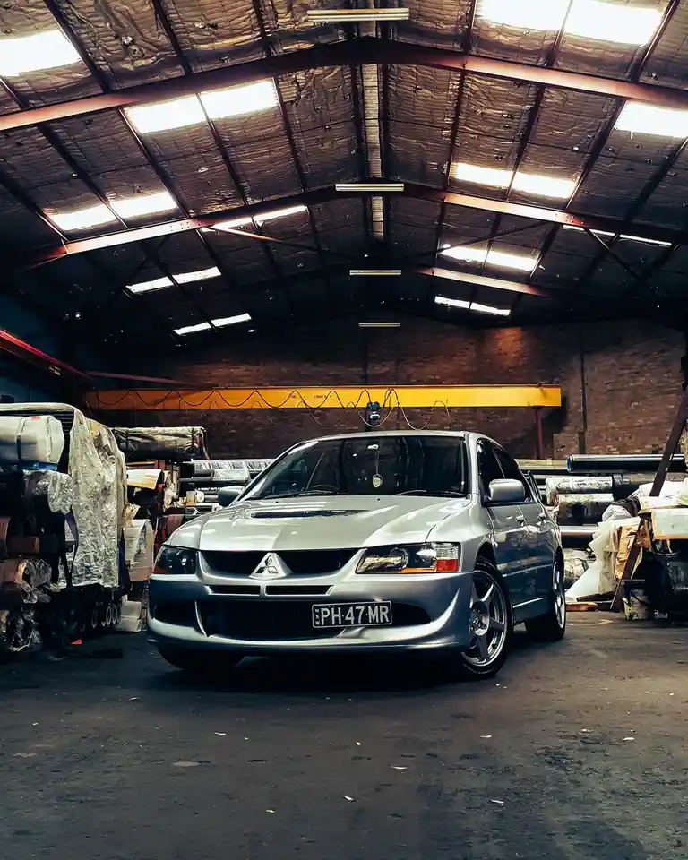 A silver Mitsubishi Lancer Evolution is parked inside a cluttered industrial warehouse with exposed metal beams and brick…
