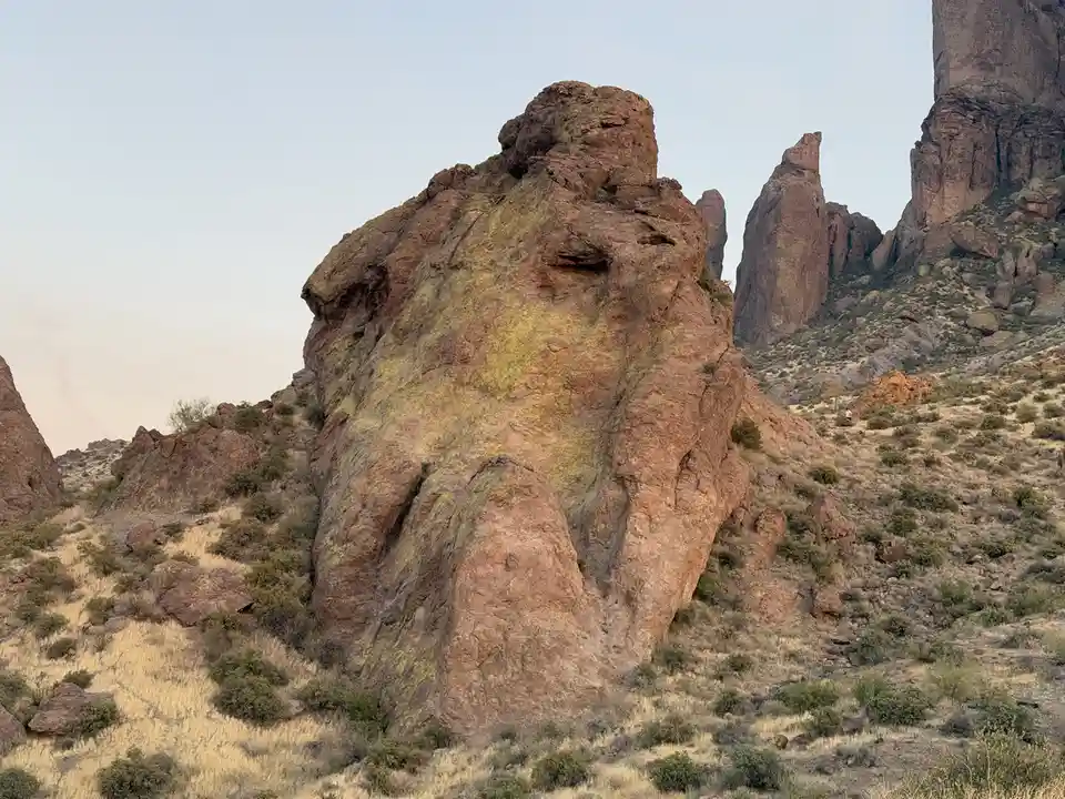 A large, reddish-brown rock formation in a desert landscape with sparse vegetation and a pale sky. The rock has a distinct…