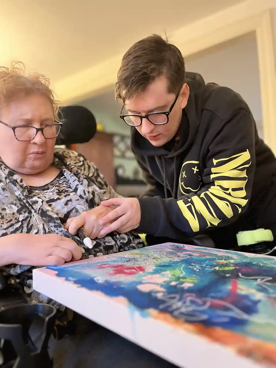 A woman in a wheelchair with glasses and a patterned top looks frustrated while holding a marker over a colorful abstract…