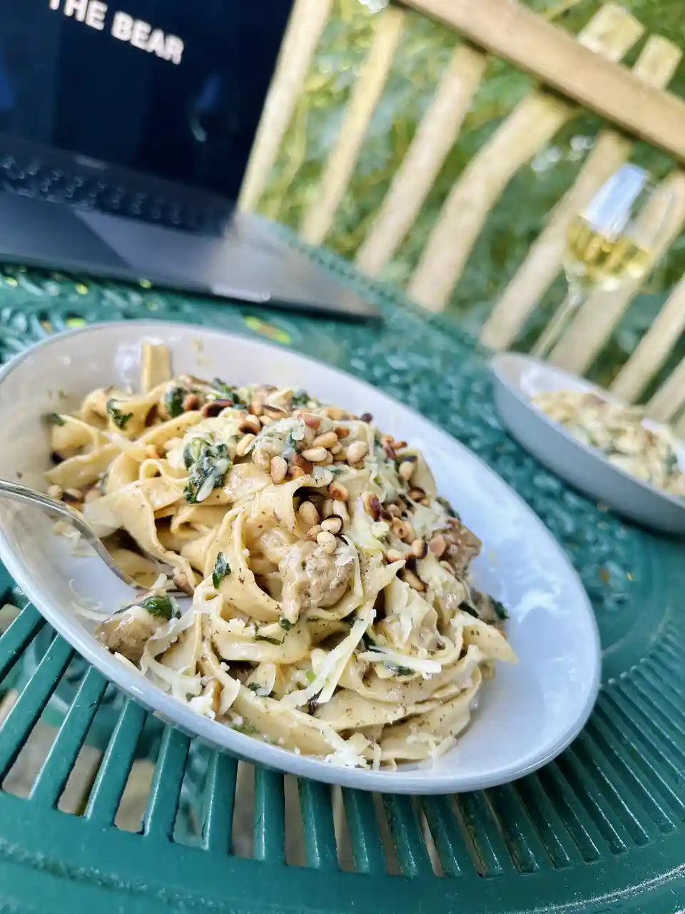 A close-up photo shows a white bowl of fettuccine pasta with pine nuts, cheese, and greens on a green outdoor table. In the…