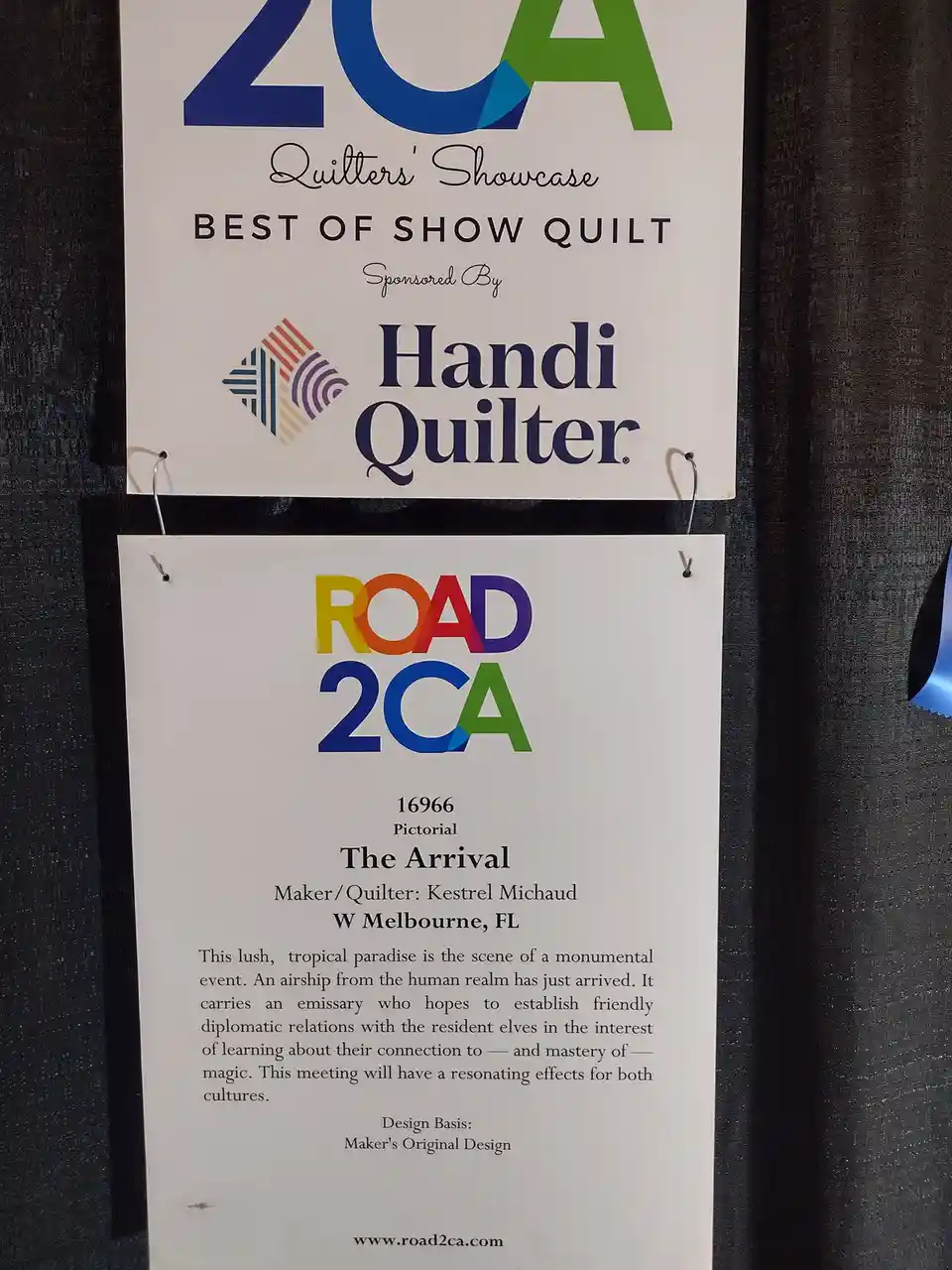 Two white signs hang on a dark curtain. The top sign reads 'ZCA Quilters' Showcase BEST OF SHOW QUILT Sponsored By Handi…