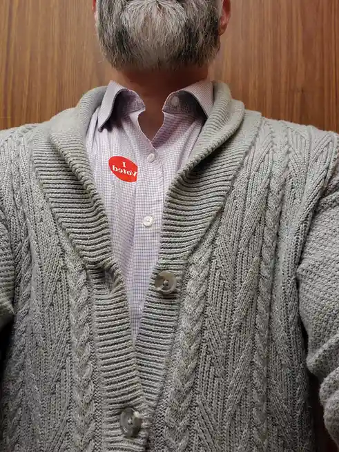 A close-up shot of a person's torso from behind, wearing a grey cable-knit cardigan over a checkered button-up shirt. A red…