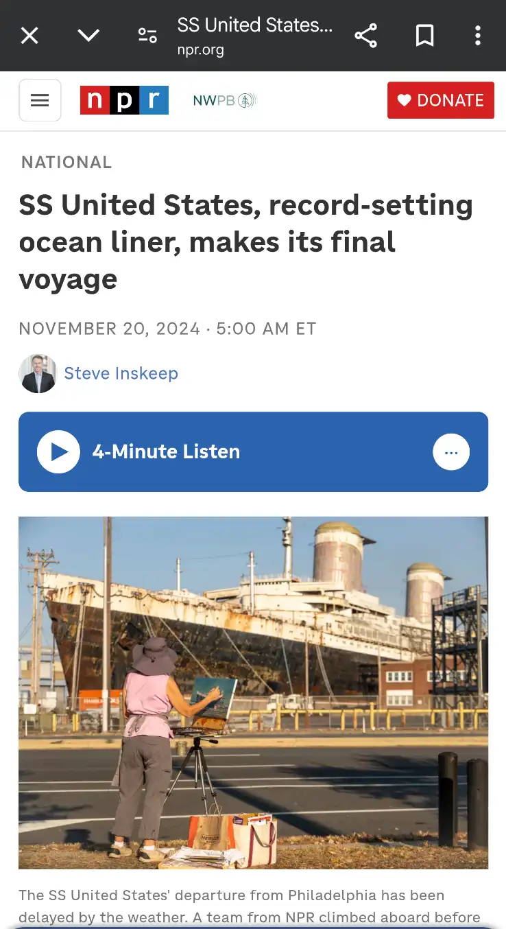A painter in a pink shirt and wide-brimmed hat stands outdoors, painting a large, rusty, decommissioned ocean liner that is…