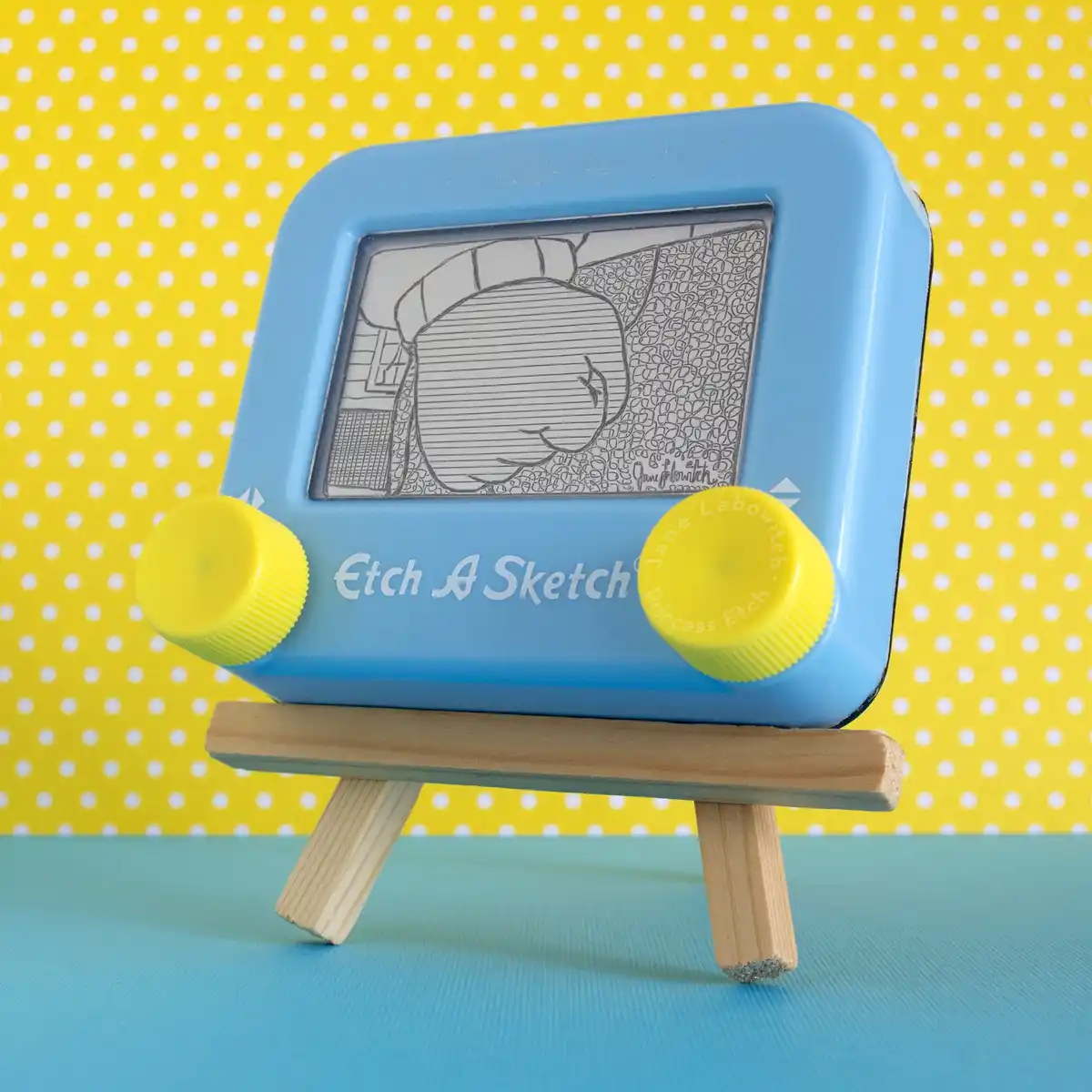 A blue Etch A Sketch toy sits on a small wooden easel against a yellow and white polka dot background. The screen displays a…
