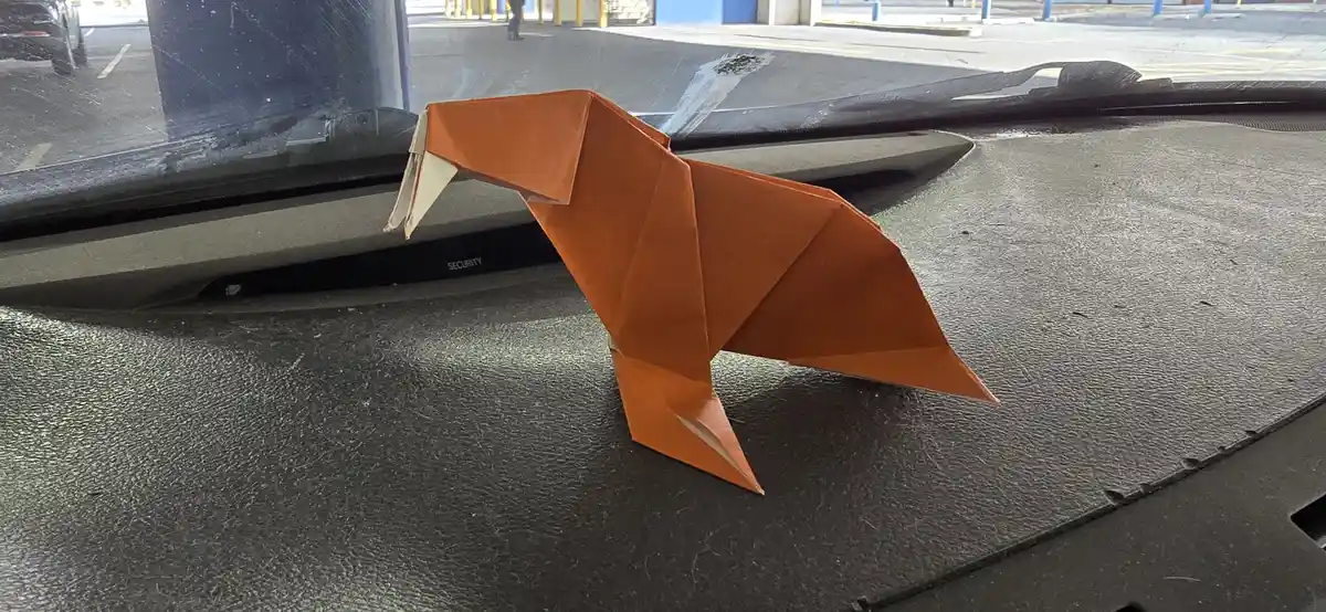 An orange origami fox sits on a car's dashboard, its head tilted as if looking out the windshield. The car's interior is…