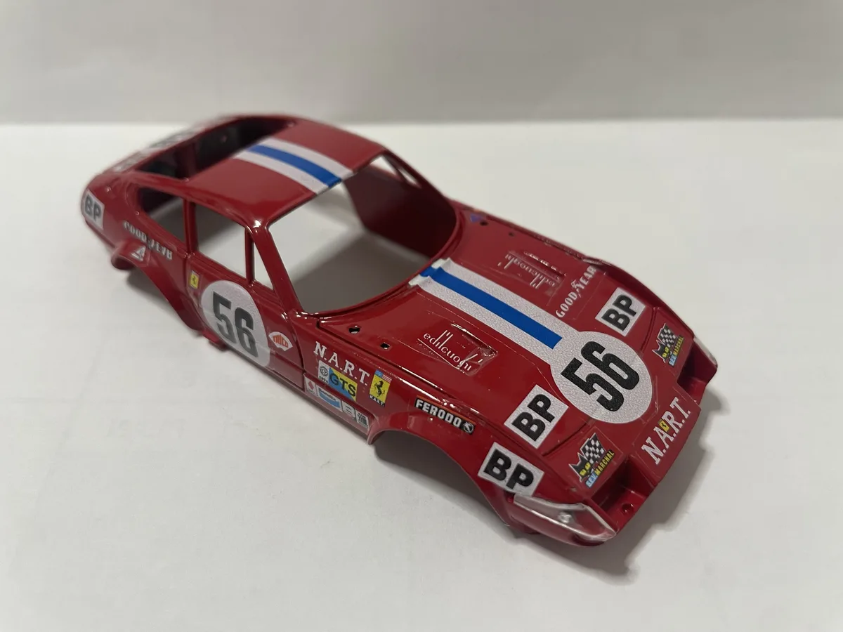 A red model race car body, number 56, with sponsor decals like BP and N.A.R.T., is shown without its interior or wheels on a…