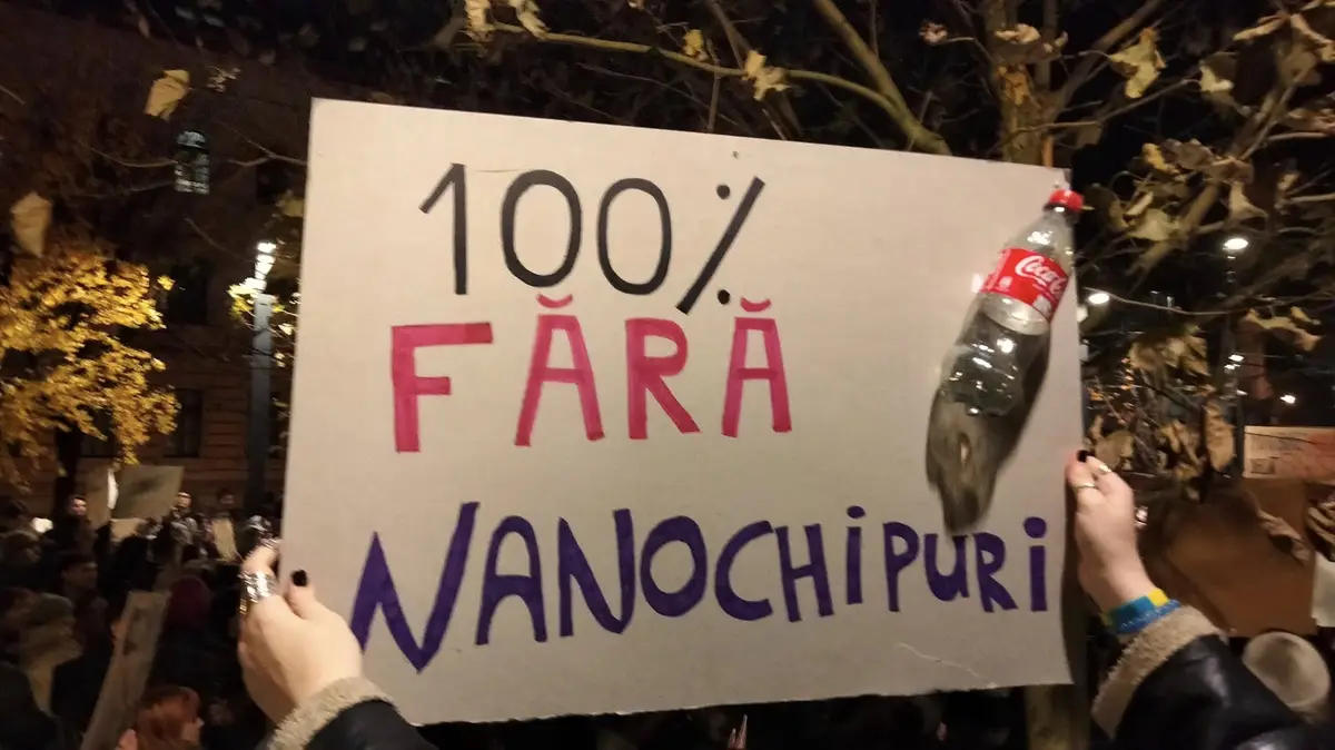 A person holds a handmade protest sign at night that reads '100% FĂRĂ NANOCHIPURI' in black, red, and purple marker. A…