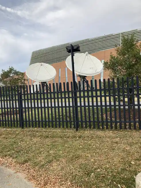 An outdoor photo shows two large white satellite dishes behind a black metal fence, with a modern building in the background…