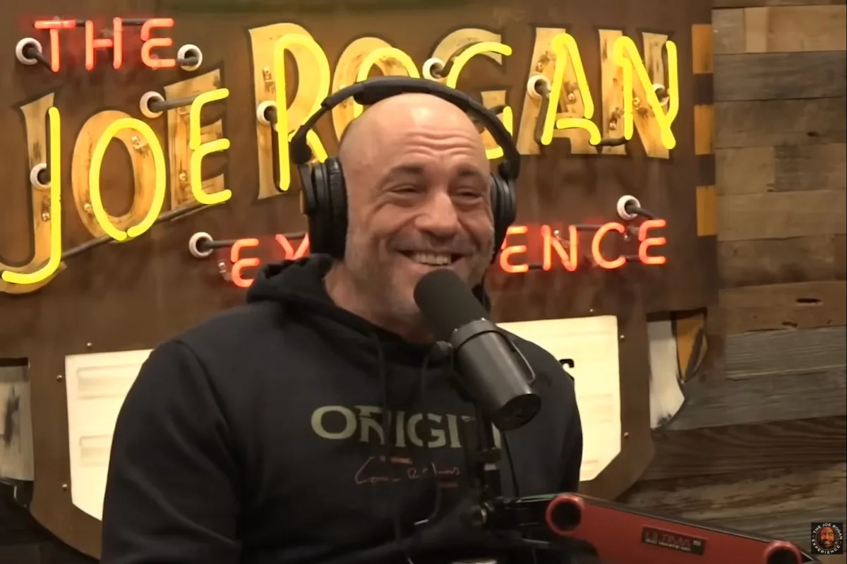 a bald man wearing headphones and a black hoodie smiles broadly while sitting in front of a microphone, with a large neon…
