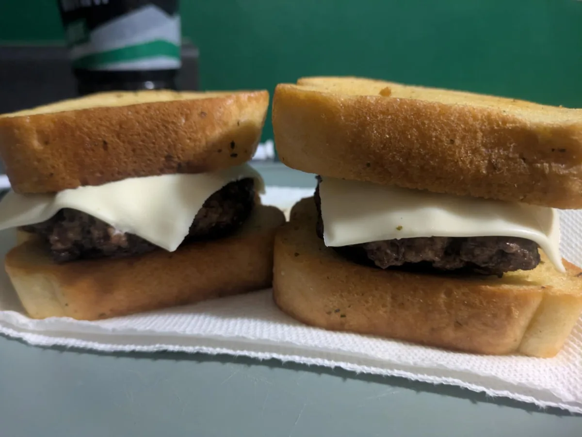 Two homemade cheeseburgers, each made with a cooked beef patty, a slice of melted white cheese, and two slices of toasted…