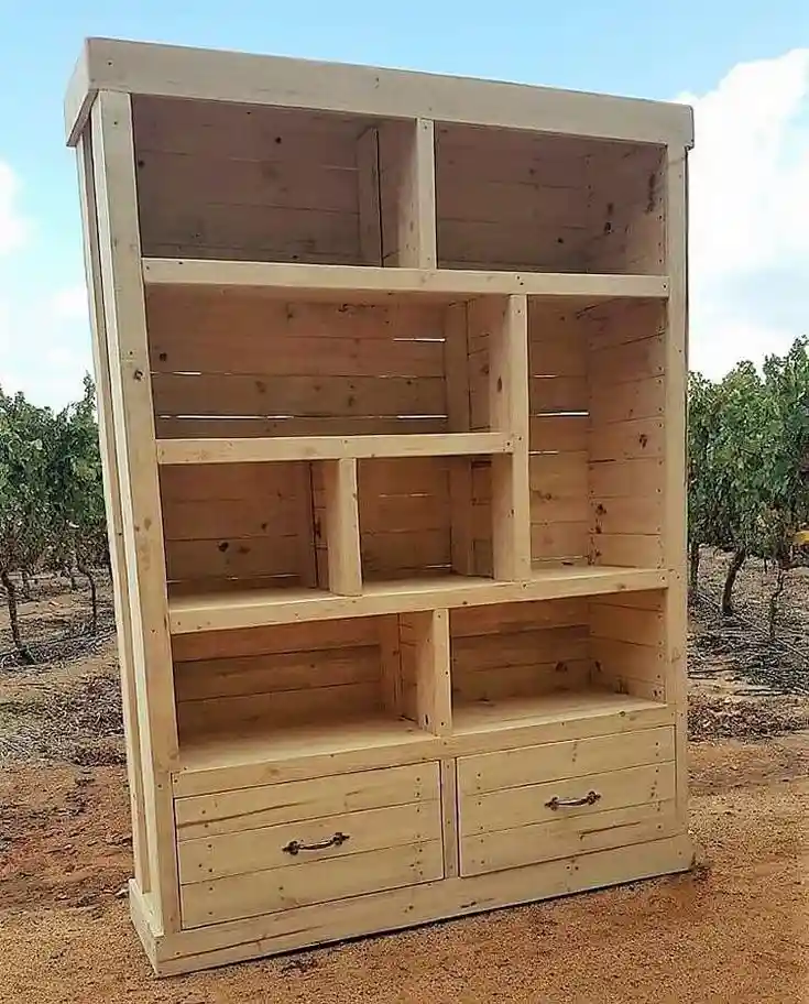 A large, unfinished wooden bookcase stands outdoors in a dry, vineyard-like setting with trees and dirt ground. The bookcase…