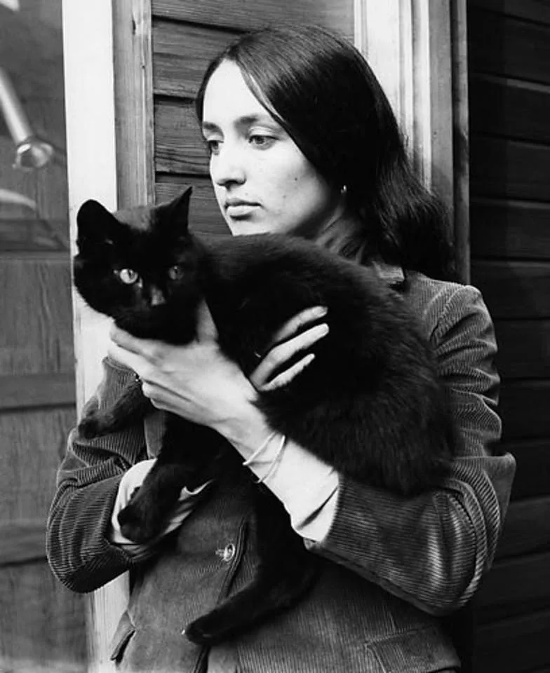A black-and-white photograph shows a woman with long dark hair holding a black cat. She is looking off to the side with a…
