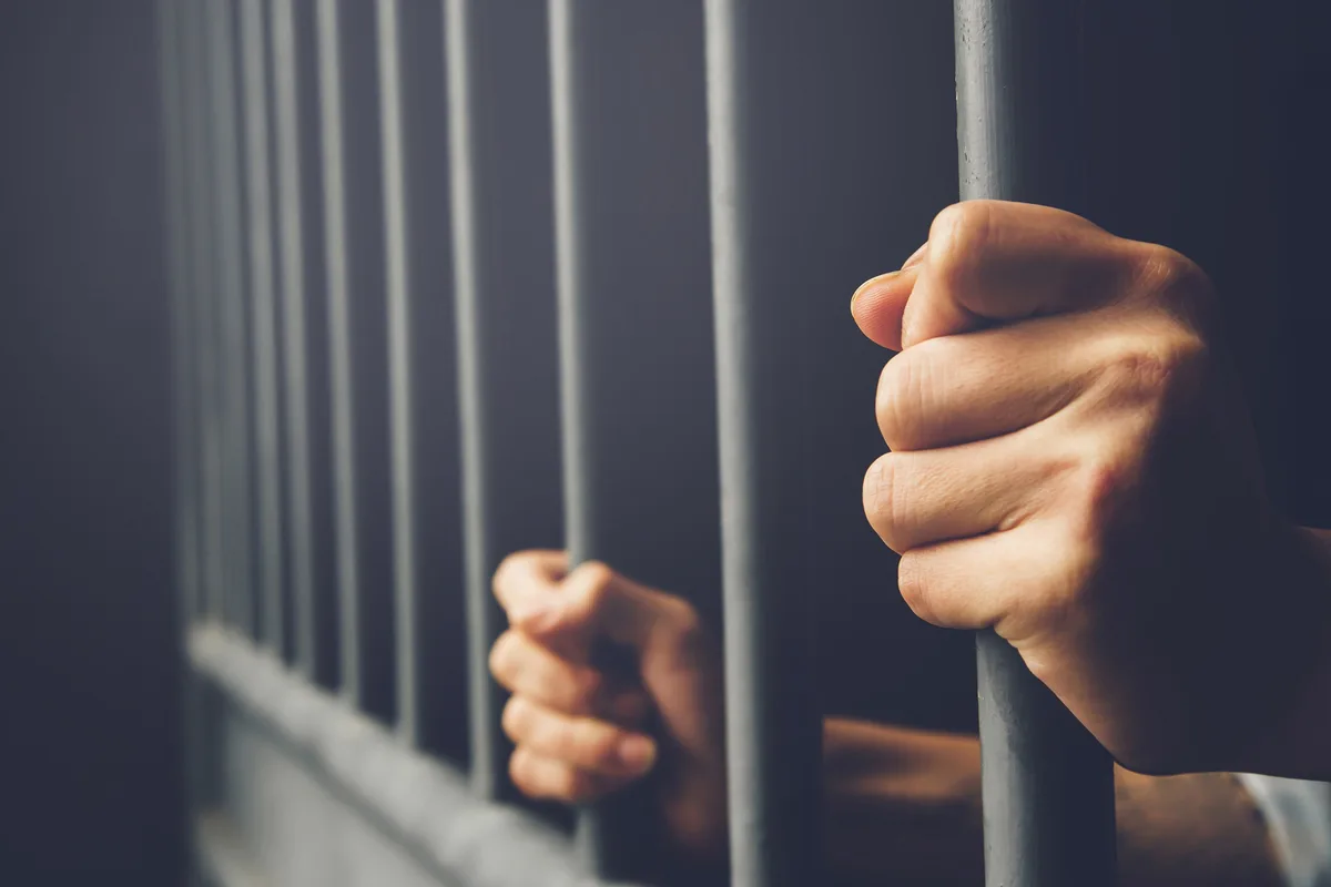 A close-up shot shows two clenched fists gripping metal prison bars, conveying confinement and frustration. The overlaid…