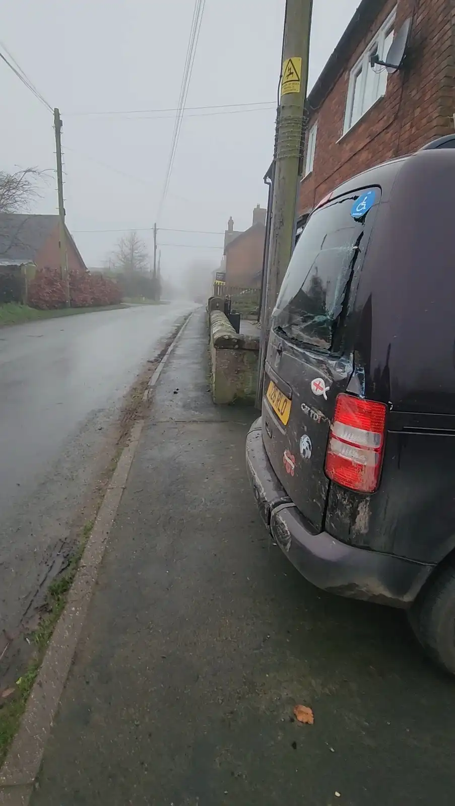A dark-colored van is parked crookedly on a wet, foggy residential street next to a brick building. The rear window is…