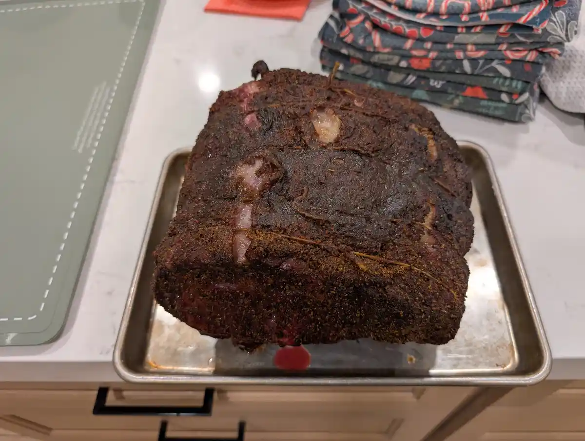 A large, heavily seasoned, dark brown meat roast sits on a metal baking tray on a white marble countertop. The meat has a…