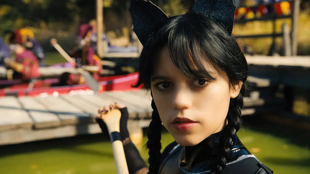 A young girl with black braided hair and black cat ears looks intensely at the camera while holding a paddle near a dock…