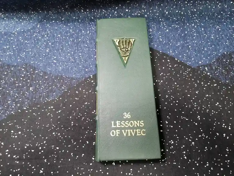 A green leather-bound book titled '36 LESSONS OF VIVEC' with a gold emblem on the cover rests on a fabric background…