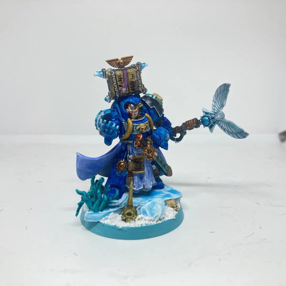 A painted miniature of a blue-armored Space Marine, likely from Warhammer 40,000, stands on a snowy base with coral and…