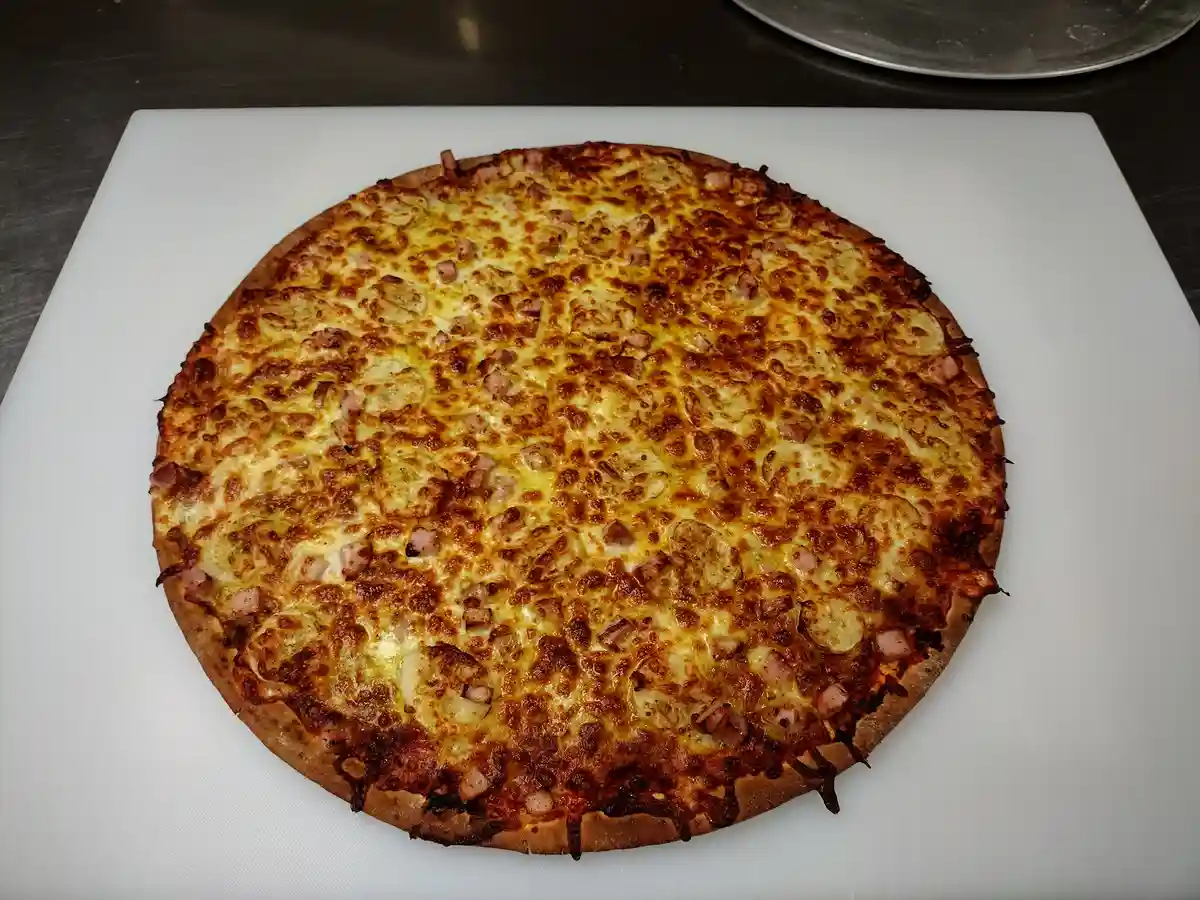 A freshly baked pizza with golden-brown cheese, ham, and potato slices sits on a white cutting board. The text overlay reads…