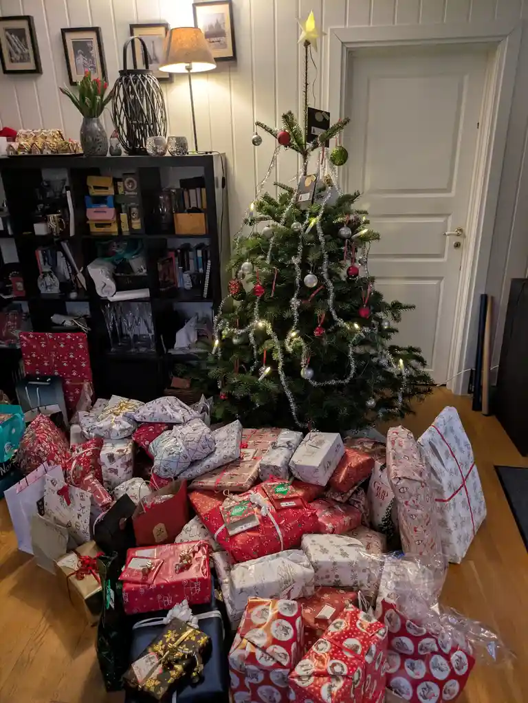 A Christmas tree decorated with lights, tinsel, and ornaments stands in a room with a large pile of wrapped gifts and…