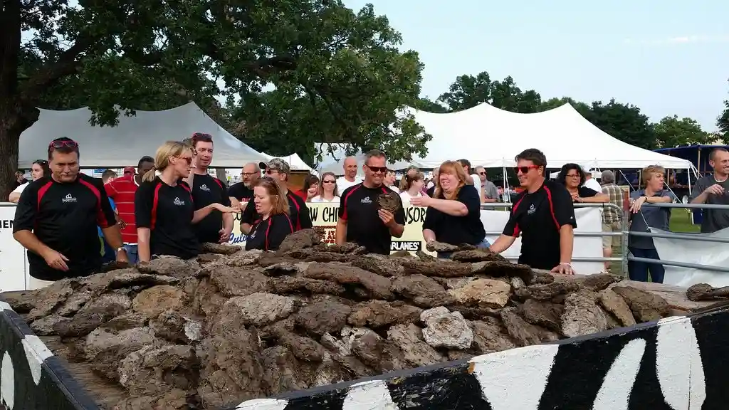 A group of people in matching black and red shirts stand behind a large pile of oyster shells at an outdoor event. One man…