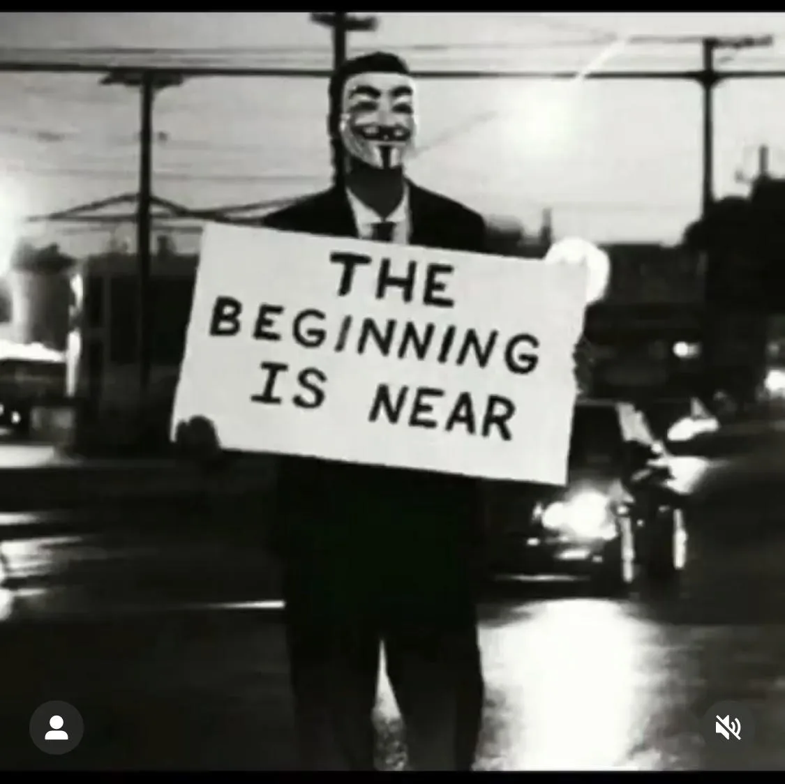 A black-and-white image shows a person wearing a Guy Fawkes mask and a suit, holding a sign that reads 'THE BEGINNING IS…