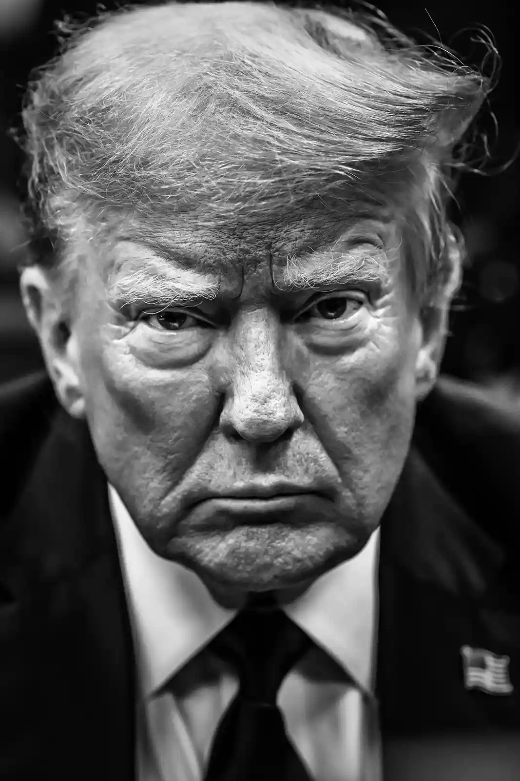 A black-and-white close-up photo of Donald Trump with a stern, displeased facial expression, wearing a suit and tie with an…