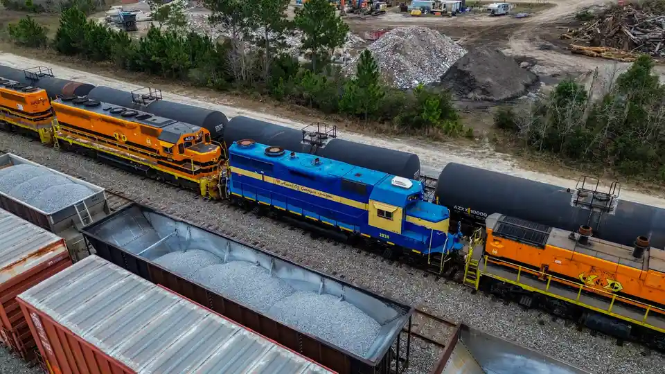 An aerial view shows multiple freight trains on parallel tracks, including a blue and yellow locomotive with the text 'i…